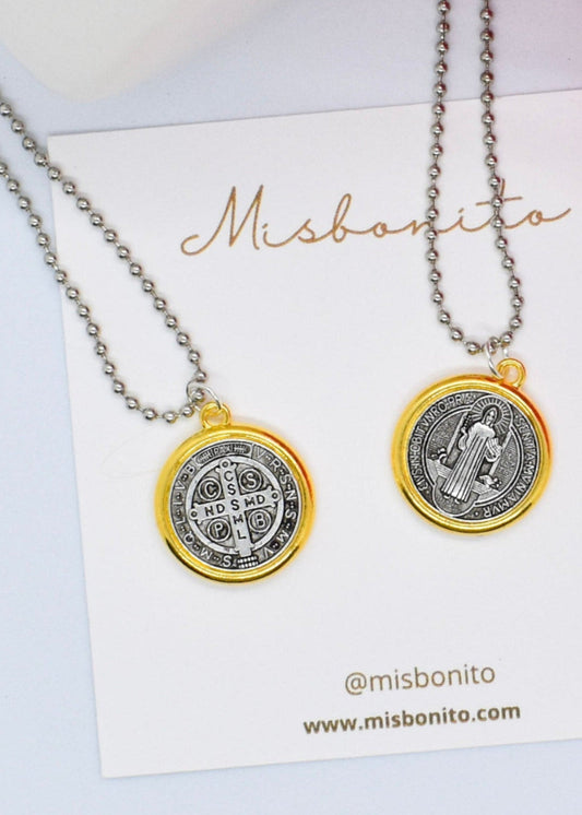 Two necklaces with round medals, one featuring a golden St Benedict pendant on a silver ball chain, two of the same necklace showing back and front of the medal. Both are displayed on a card with the brand name 'Misbonito'.