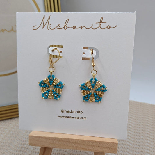 A pair of gold and turquoise beaded dangle earrings displayed on a white Misbonito card stand, which is placed on a small wooden easel.