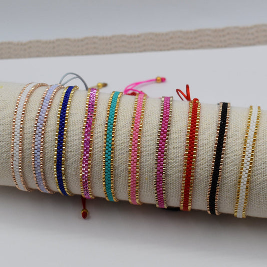 A collection of colorful woven cord bracelets displayed on a beige fabric-covered cylindrical stand, resting on a white card over a beige textured mat.