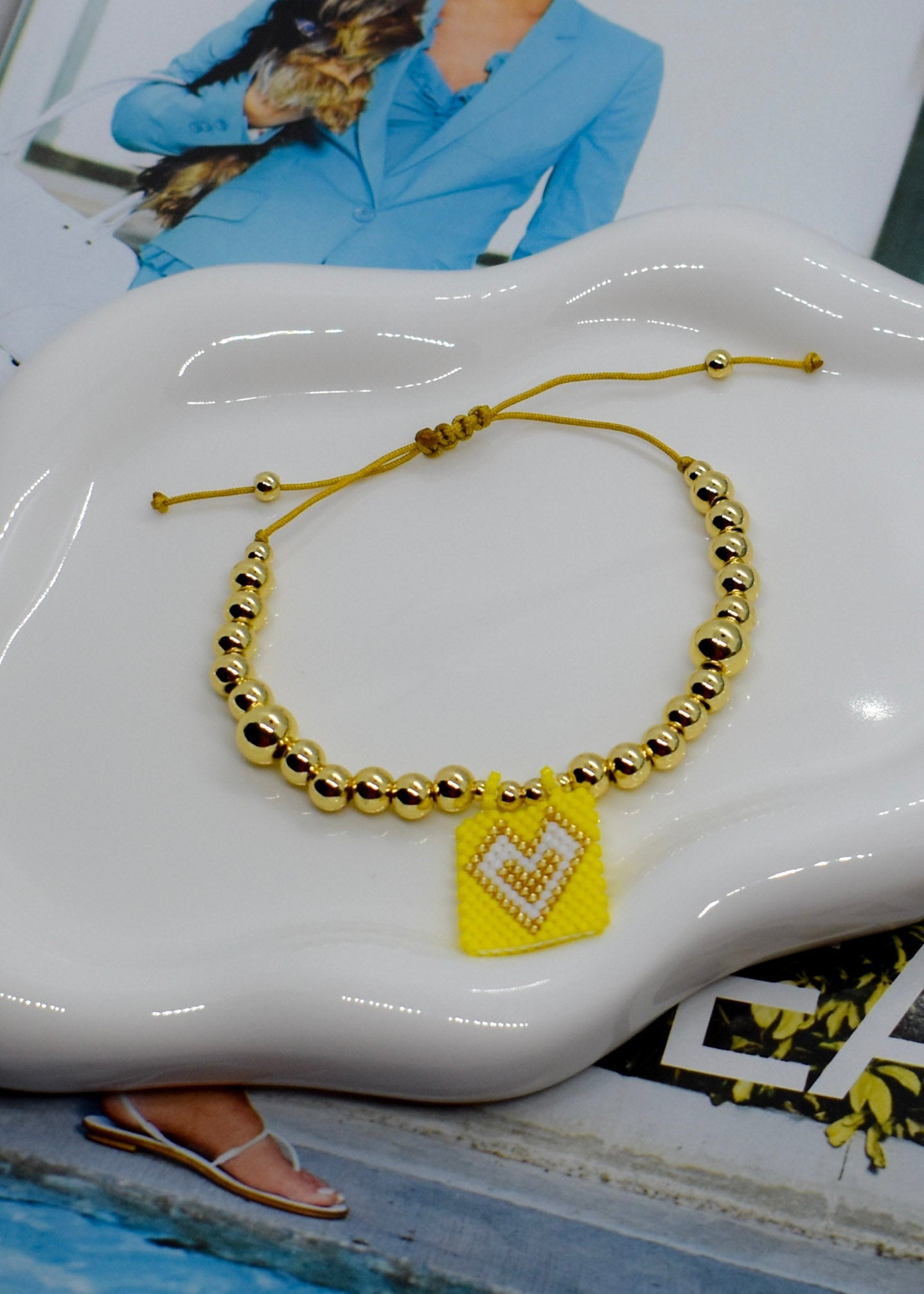 Gold beaded bracelet with a yellow heart charm on a white dish.