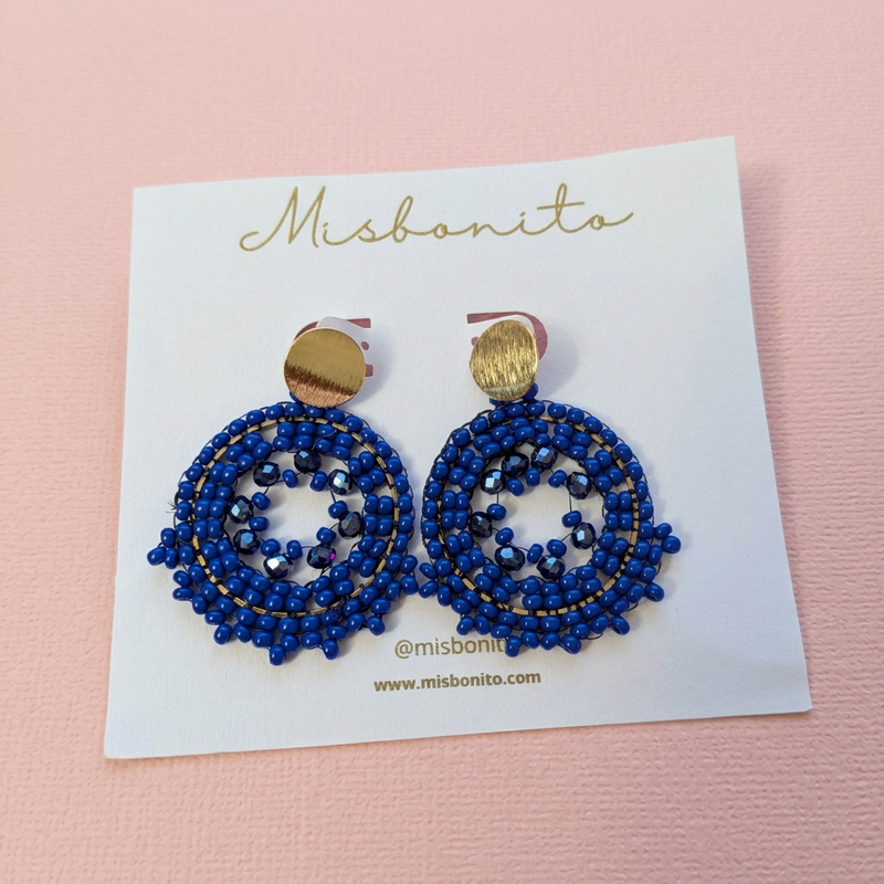 A pair of Misbonita beaded drop earrings with gold-tone tops and blue seed beads, displayed on a white brand card on a wooden surface.