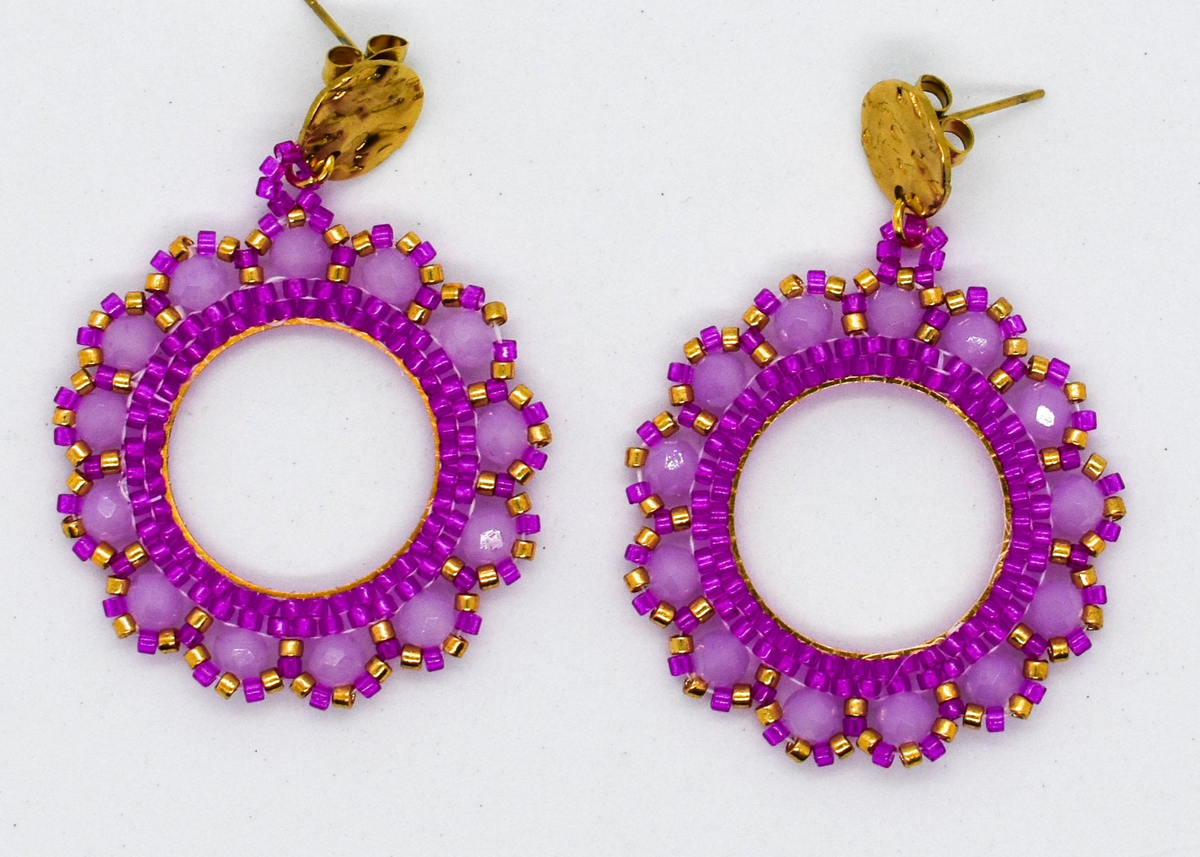 A pair of handmade circular drop earrings with deep red beaded rings and gold-tone fishhook wires, displayed on a light gray surface with blurred pink and yellow flowers in the background.