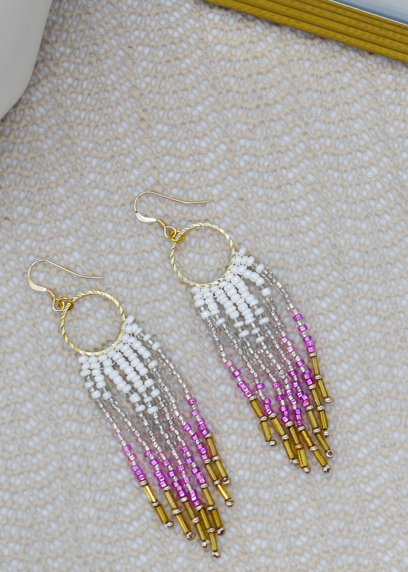 Beaded earrings with white, pink, and gold beads on a textured surface.