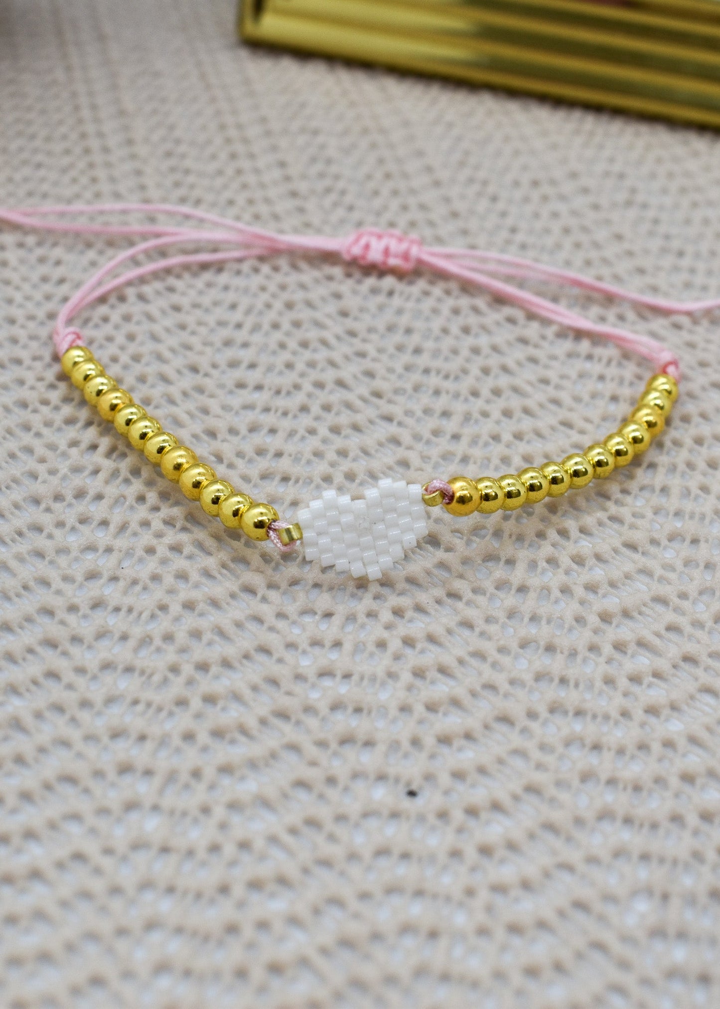 Bracelet with gold beads and a white charm on a pink string against a textured beige background