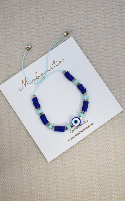 A beaded bracelet with royal blue and light blue beads, a central evil eye charm, and small gold spacer beads, displayed on a white Misshanti brand card over a beige textured fabric background.