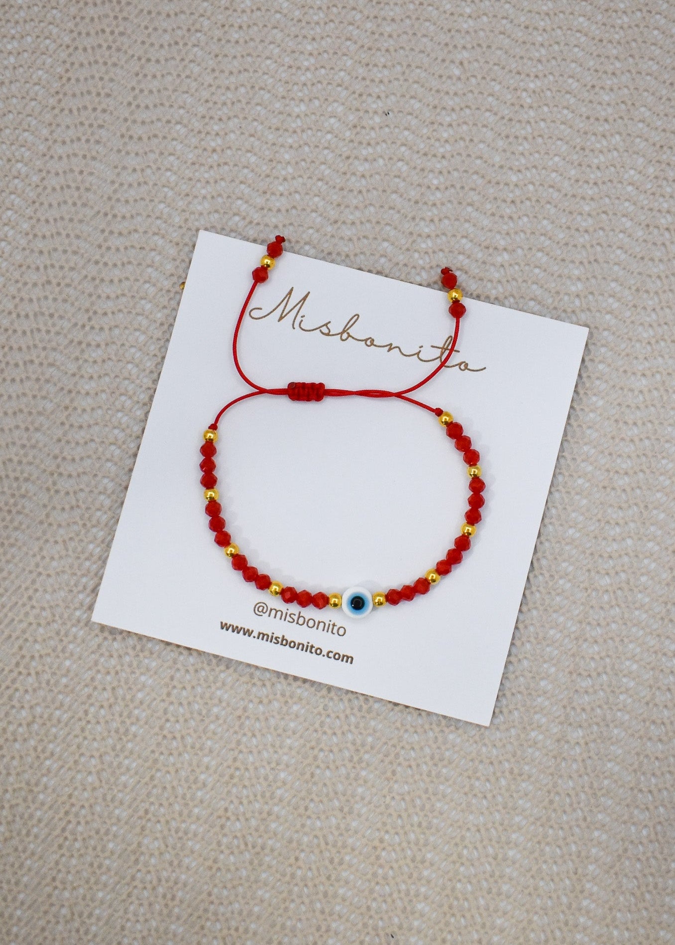 A red beaded bracelet with small gold spacer beads and a central blue evil eye charm, displayed on a white branded card over a beige textured fabric background.