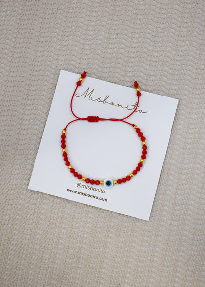 A red beaded bracelet with small gold spacer beads and a central blue evil eye charm, displayed on a white branded card over a beige textured fabric background.