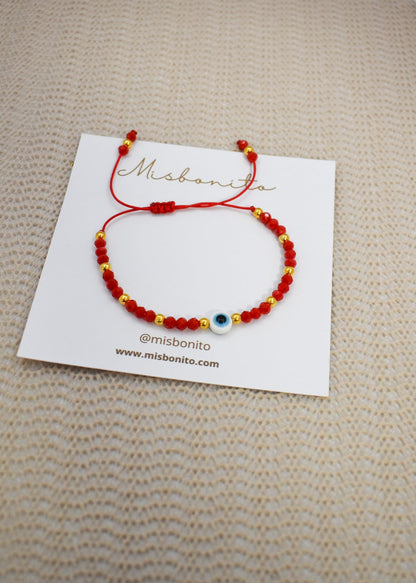 A red beaded cord bracelet with a blue and white evil eye charm, displayed on a white branded card over a beige textured fabric background.