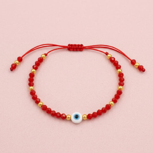 A red beaded cord bracelet with a blue and white evil eye charm, displayed on a white branded card over a beige textured fabric background.