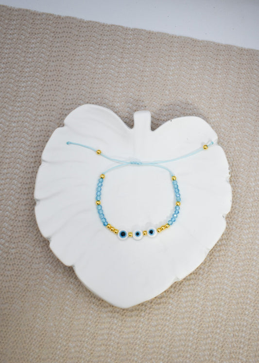 A delicate beaded bracelet with light blue cord, light blue beads, and gold spacer beads, featuring a central evil eye motif. The bracelet is displayed on a white ceramic leaf-shaped dish over a beige woven fabric background.