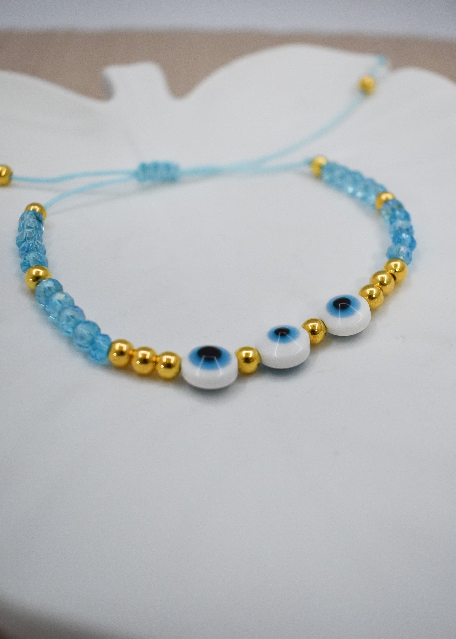 A beaded bracelet with light blue beads, shiny gold spacer beads, and three white evil eye beads, displayed on a white surface.