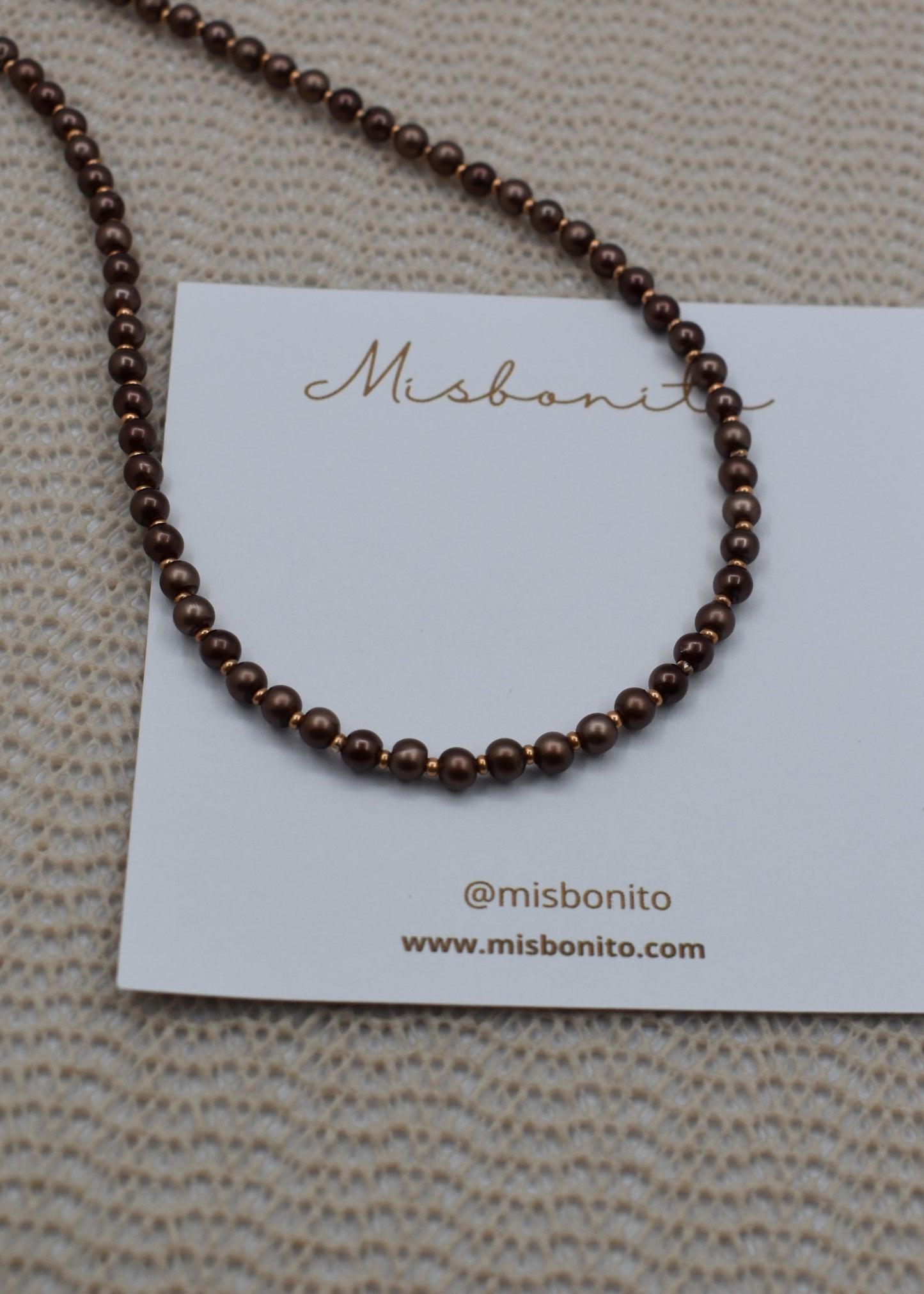A dark brown beaded necklace displayed on a white Misbonito brand card, set against a textured beige woven background.