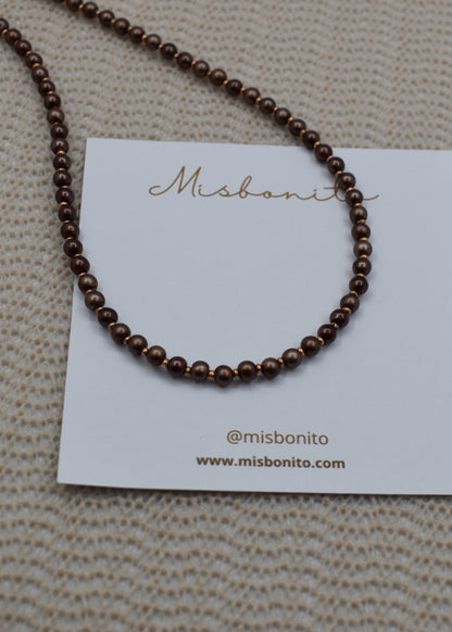 A dark brown beaded necklace displayed on a white Misbonito brand card, set against a textured beige woven background.