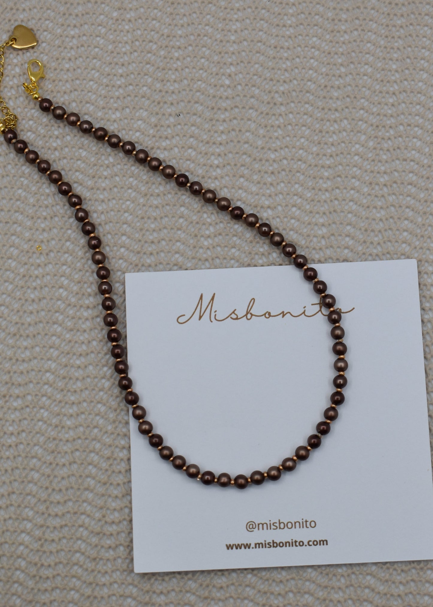 A dark brown beaded necklace with gold-toned hardware, displayed on a white brand card against a textured beige woven background.