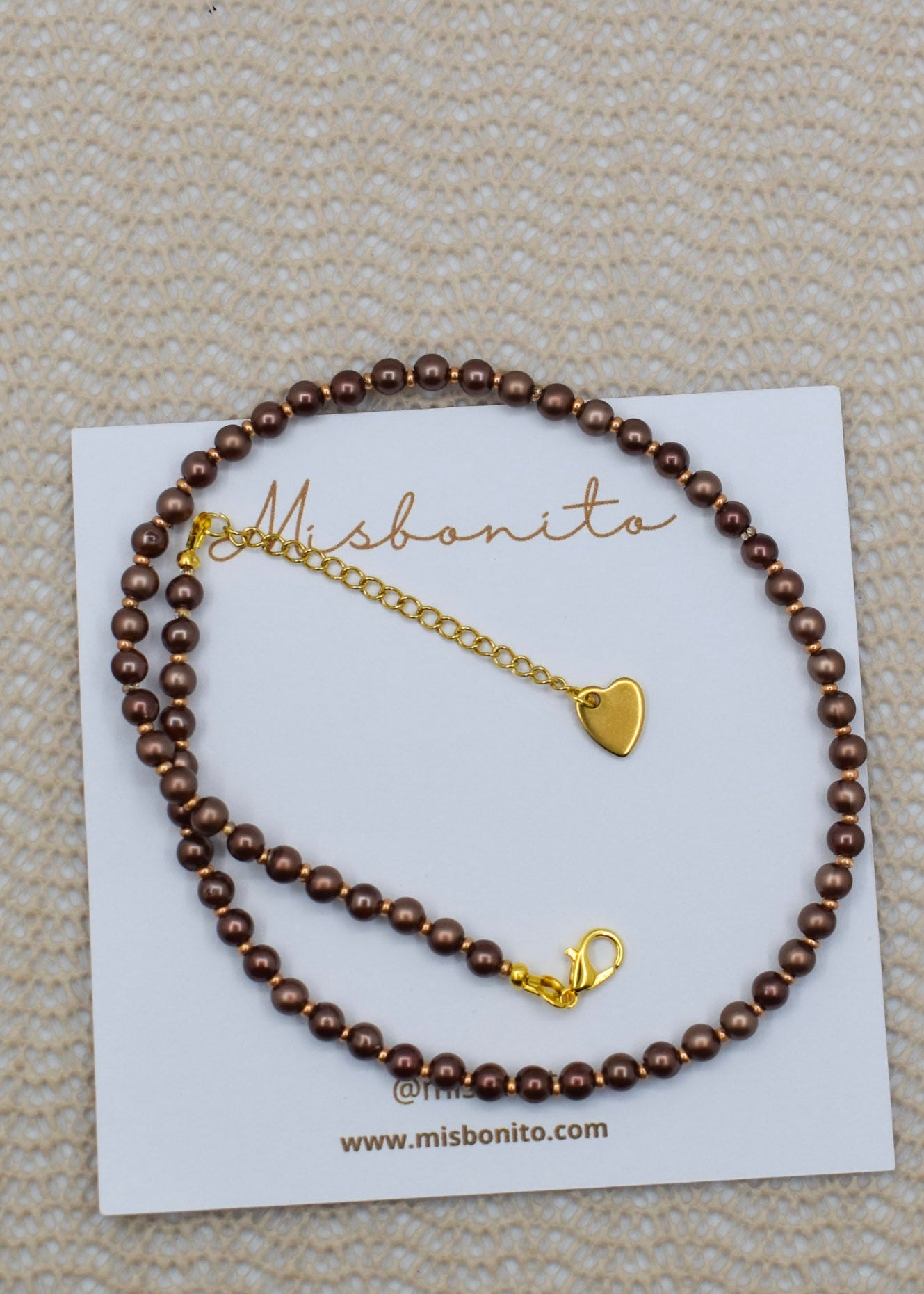 A brown beaded bracelet with a gold heart charm and lobster clasp, displayed on a white Misbonito brand card over a textured beige fabric background.