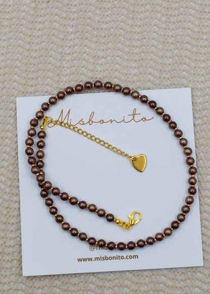 A brown beaded bracelet with a gold heart charm and lobster clasp, displayed on a white Misbonito brand card over a textured beige fabric background.
