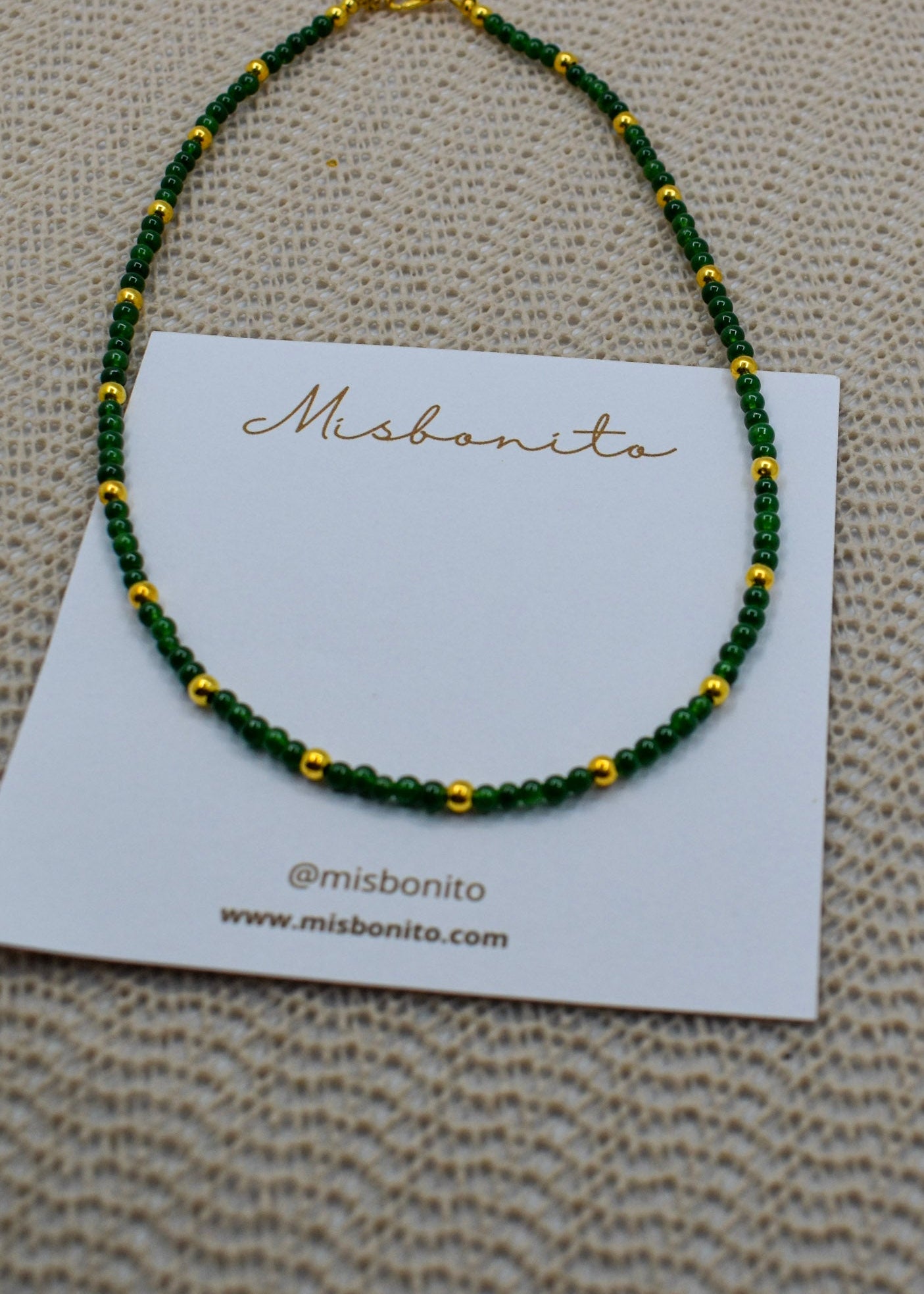 A beaded necklace with dark green and gold beads displayed on a white card labeled Misbonito, set against a textured beige fabric background.