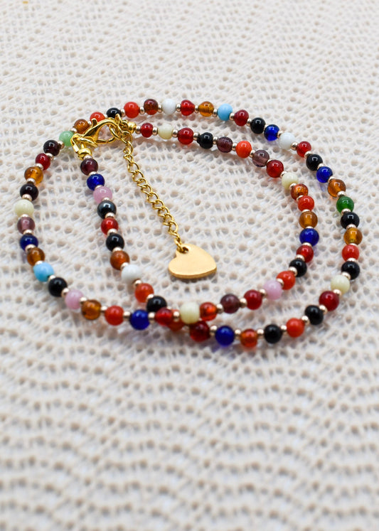 A beaded necklace with multicolor round beads and a gold heart charm, displayed on a textured white fabric background.