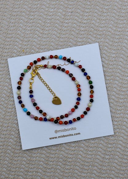 A beaded bracelet with multicolor round beads and a gold heart charm, displayed on a white branded card over a beige textured fabric background.