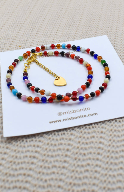 A beaded double-strand necklace with colorful round beads and a gold heart charm, displayed on a white card over a beige textured knit surface.