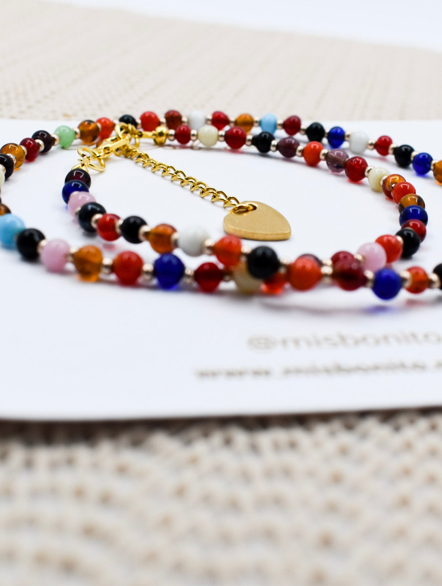 A close-up of a beaded bracelet with multicolored round beads and a gold heart charm, resting on a white card over a textured beige fabric background.