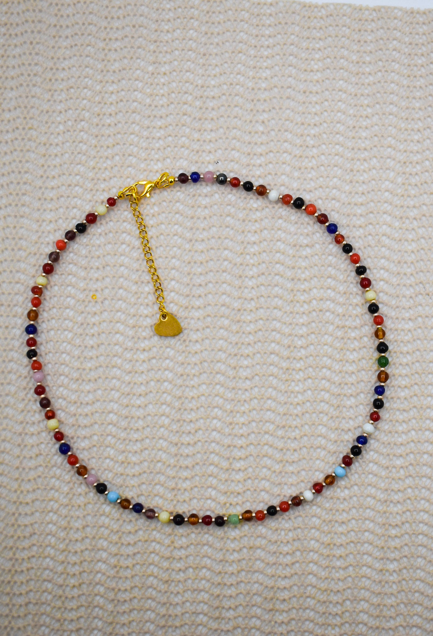 A beaded necklace with multicolor round beads and a gold heart charm, displayed on a textured cream woven fabric background.