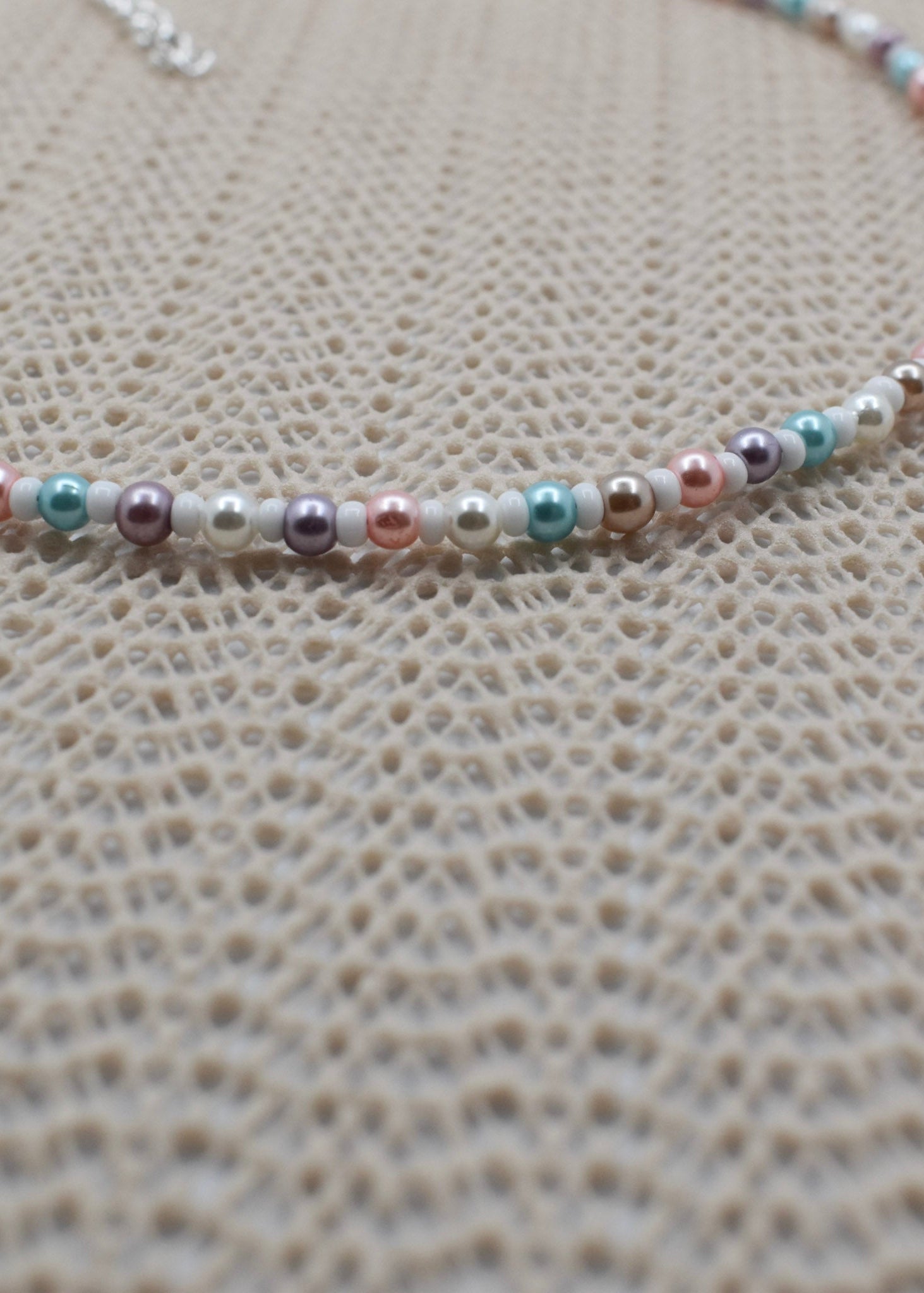 A close-up of a beaded necklace featuring small pearls in pastel pink, mint green, lavender, and white, laid out on a textured beige woven surface.