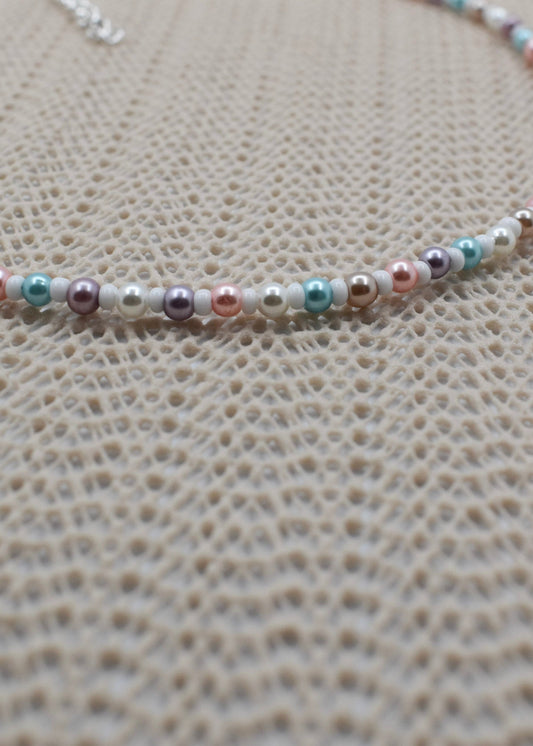 A close-up of a beaded necklace featuring small pearls in pastel pink, mint green, lavender, and white, laid out on a textured beige woven surface.
