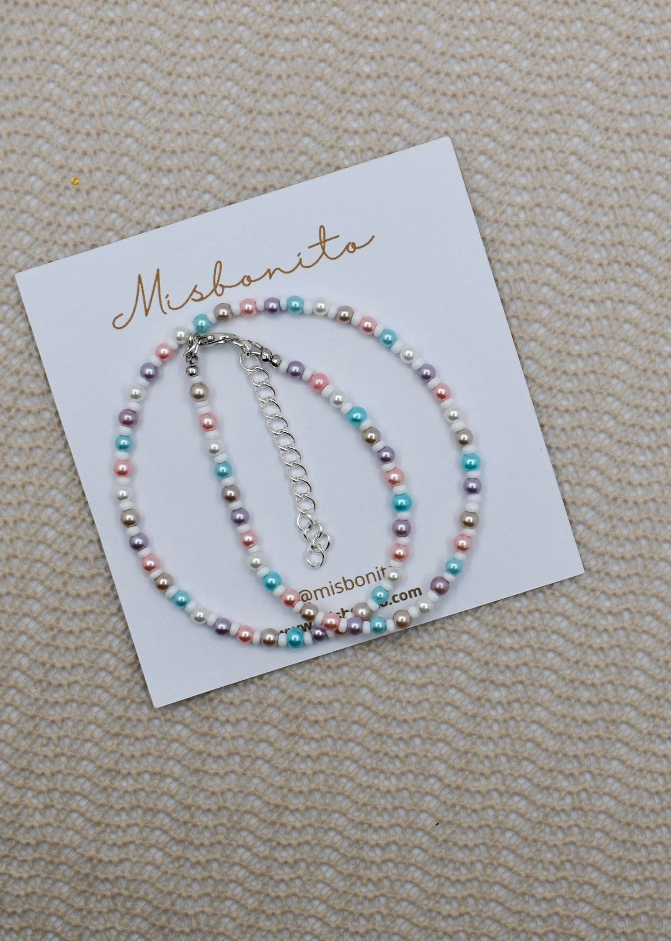A beaded bracelet with pastel pink, blue, purple, and white pearls on a white display card branded Misbanita, placed on a textured beige fabric background.