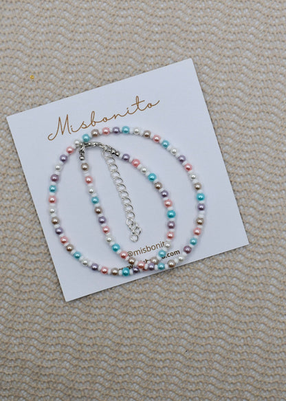 A beaded bracelet with pastel pink, blue, purple, and white pearls on a white display card branded Misbanita, placed on a textured beige fabric background.