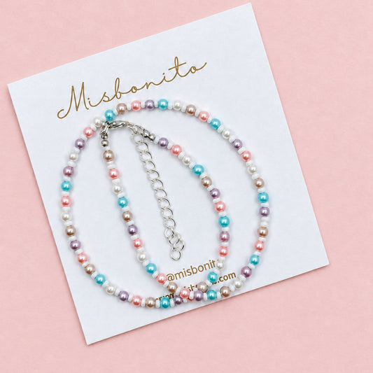 A beaded bracelet with pastel pink, blue, purple, and white pearls on a white display card branded Misbanita, placed on a textured beige fabric background.