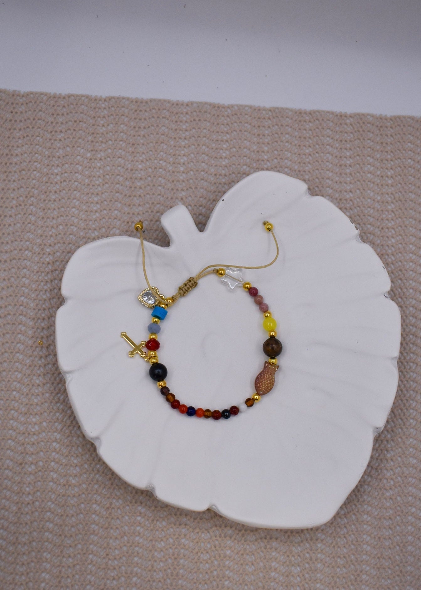 A delicate gold-tone beaded bracelet with colorful gemstone-like beads and a small cross charm, displayed on a white leaf-shaped ceramic dish over a beige textured surface.