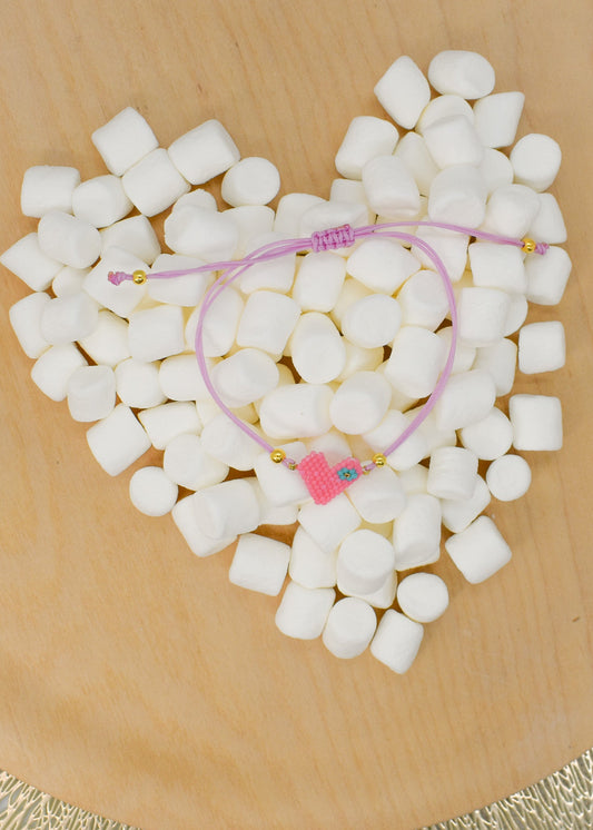 Pink bracelet with a heart charm on a heart-shaped arrangement of white marshmallows on a wooden surface.