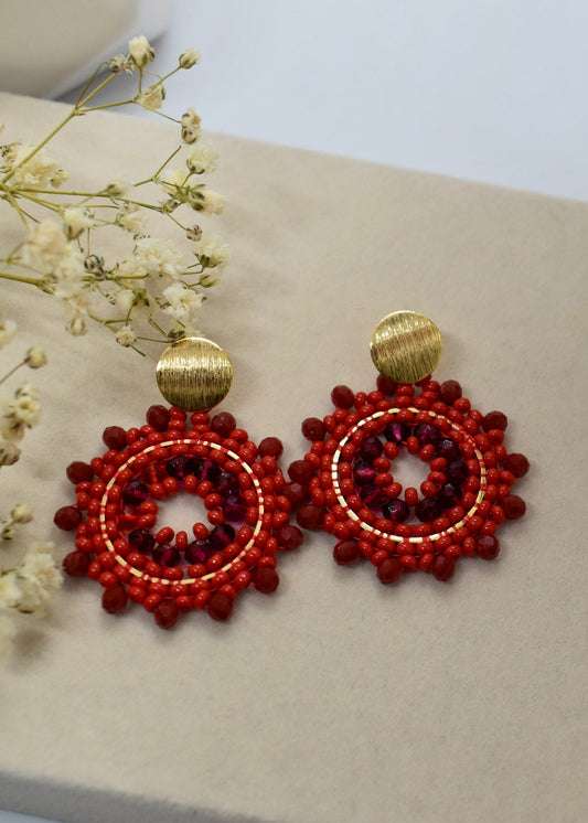 Red beaded earrings with gold accents on a beige surface with white flowers.