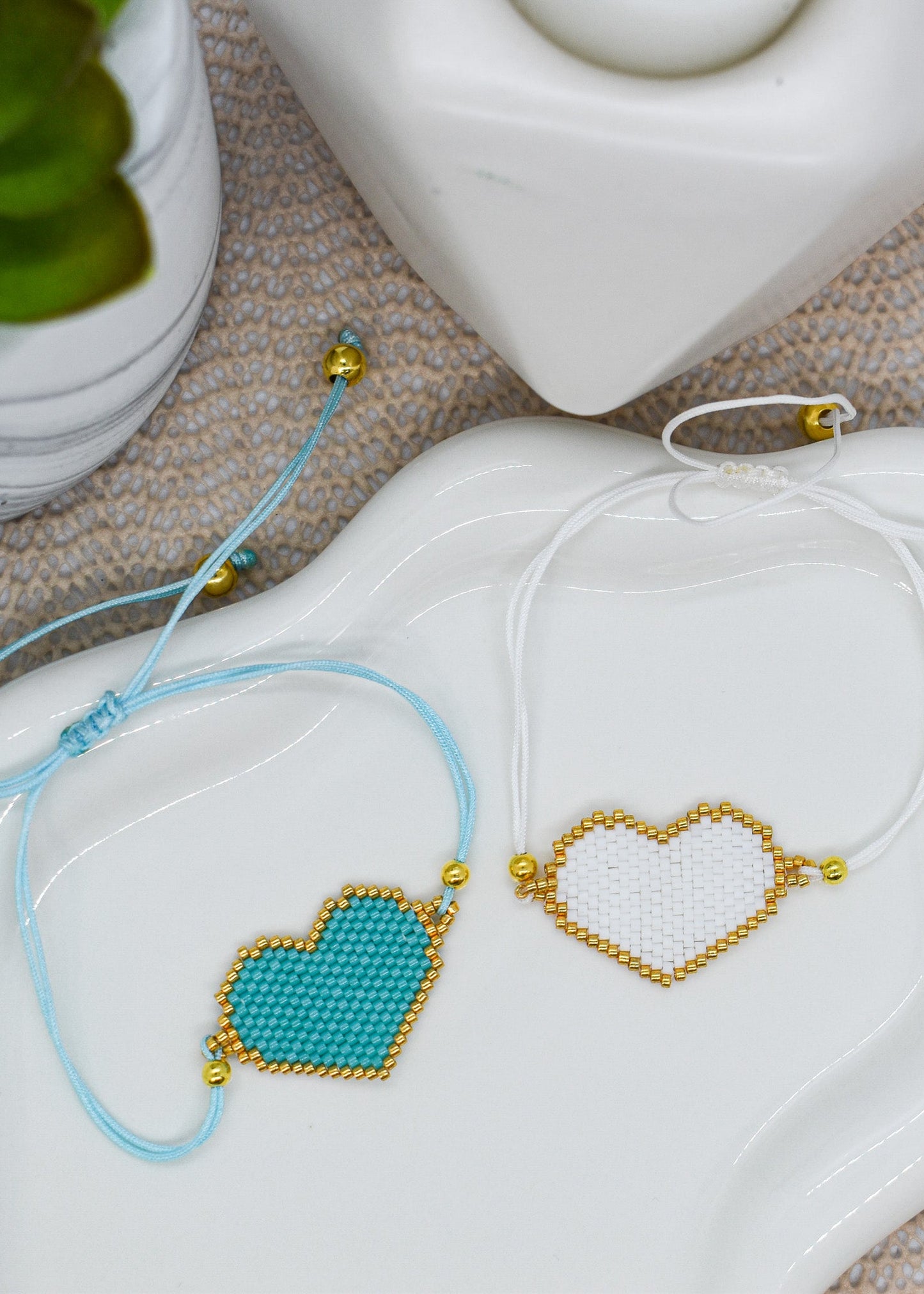 Two heart-shaped beaded bracelets on a white dish with a plant in the background.