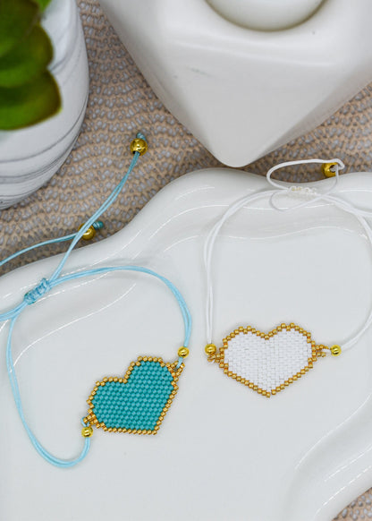 Two heart-shaped beaded bracelets on a white dish with a plant in the background.