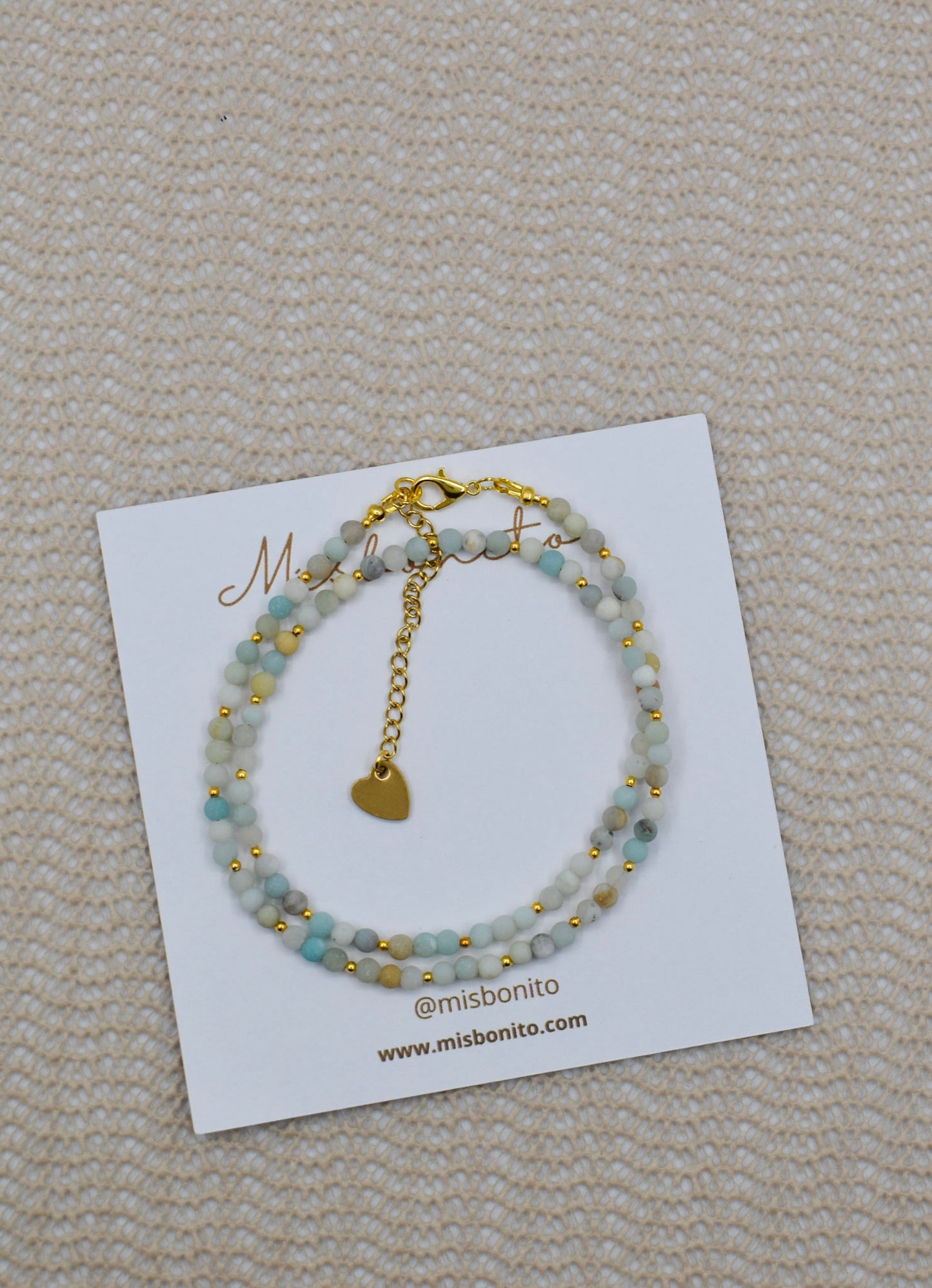 A double-strand beaded bracelet with light blue-green beads and gold-tone details, displayed on a white Missoni card on a beige textured fabric background.