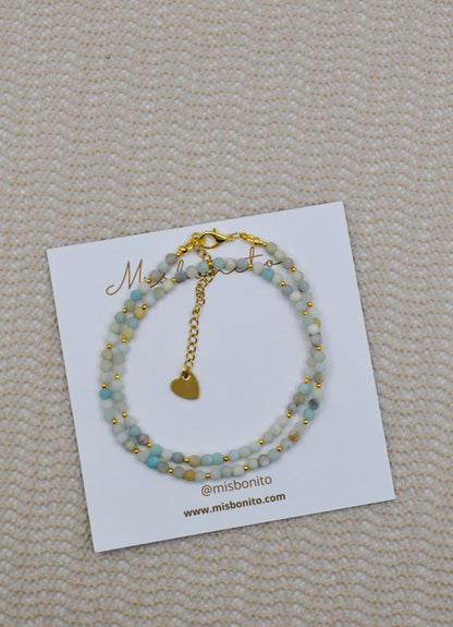 A double-strand beaded bracelet with light blue-green beads and gold-tone details, displayed on a white Missoni card on a beige textured fabric background.