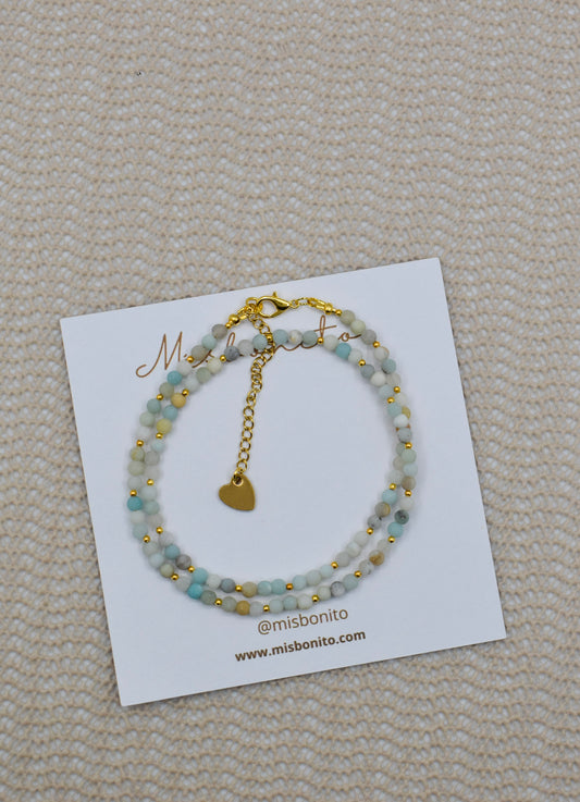 A double-strand beaded bracelet with light blue-green beads and gold-tone details, displayed on a white Missoni card on a beige textured fabric background.