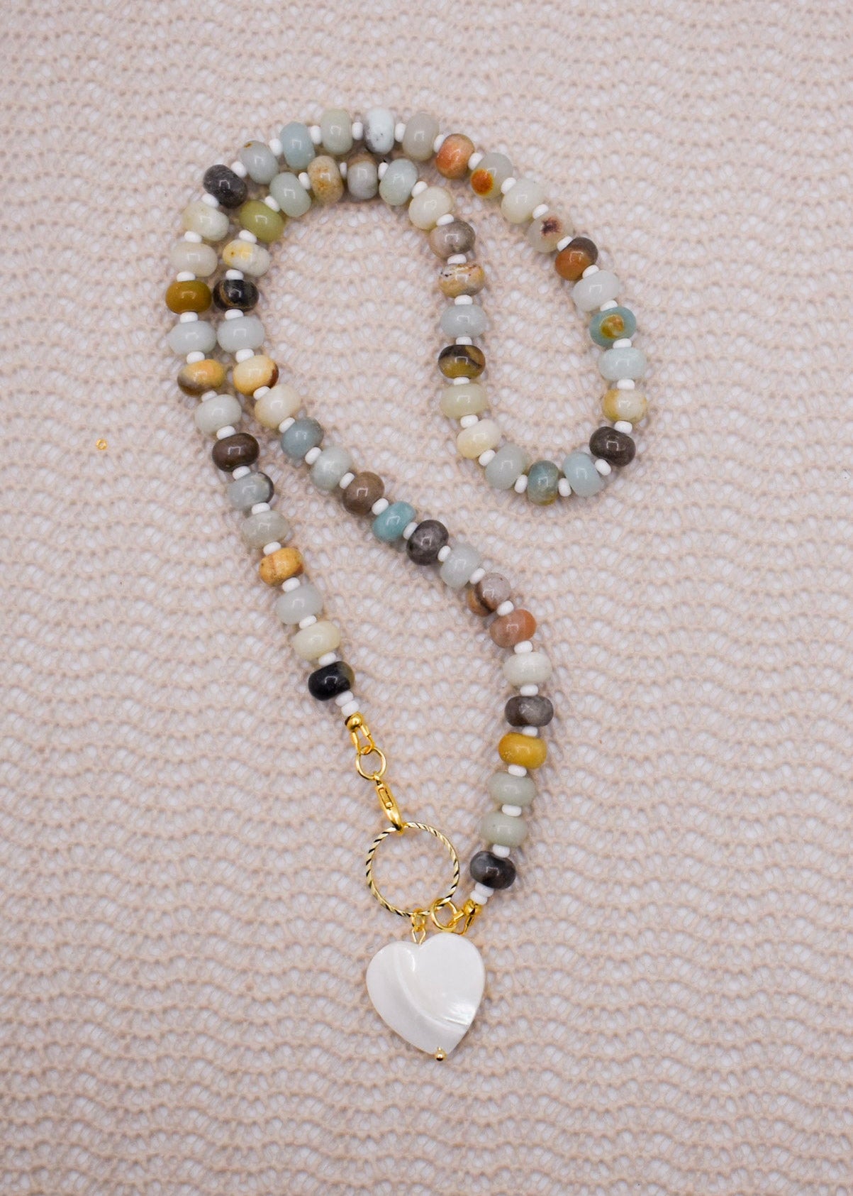A beaded necklace with amazonite necklace in shades of gray, brown, yellow, and blue, featuring a gold-tone clasp and a white heart-shaped pendant, laid flat on a textured light pink fabric background.