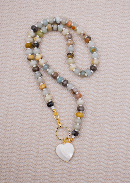 A beaded necklace with amazonite necklace in shades of gray, brown, yellow, and blue, featuring a gold-tone clasp and a white heart-shaped pendant, laid flat on a textured light pink fabric background.