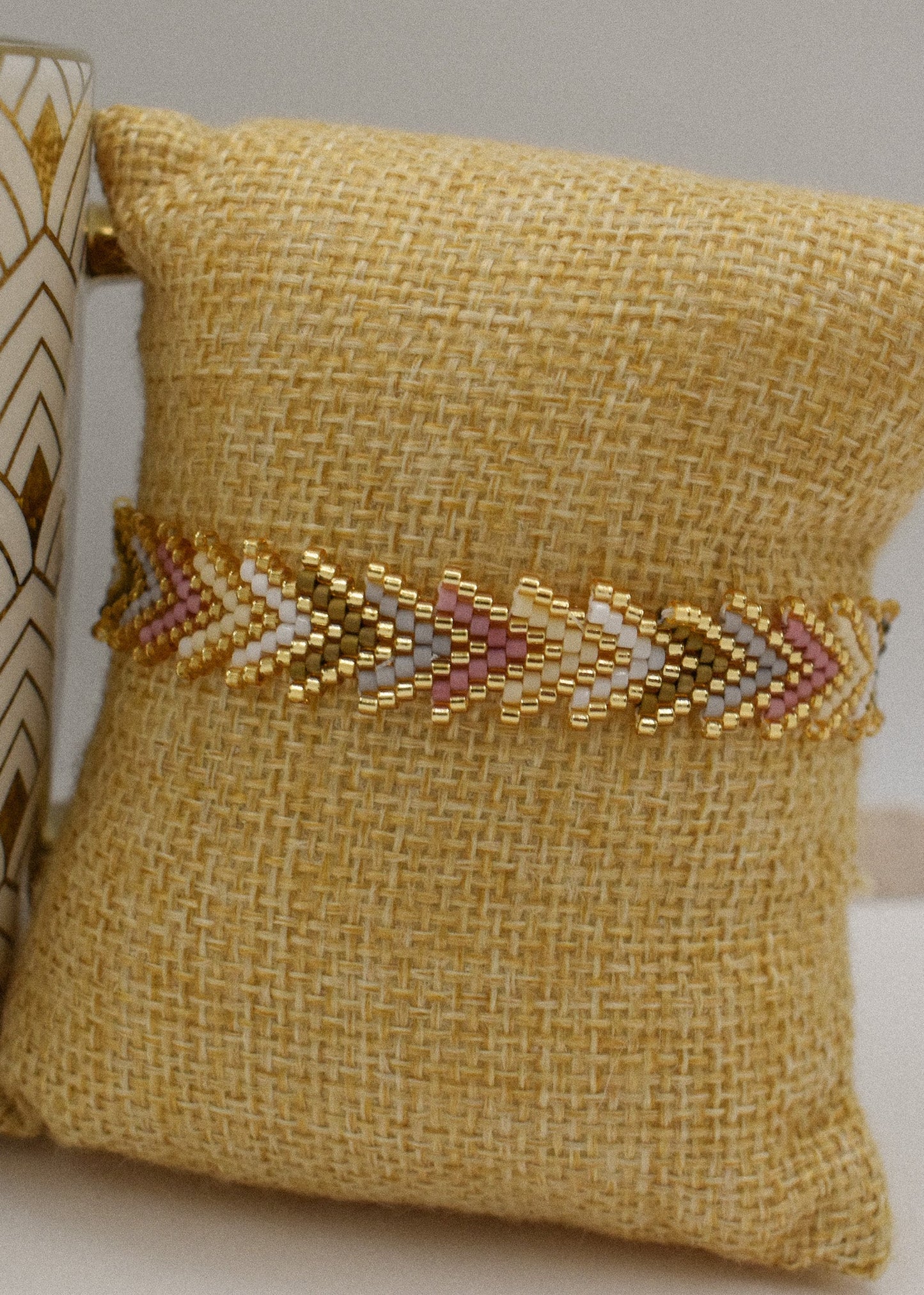 A gold beaded chevron-patterned bracelet displayed on a yellow burlap pillow, with a decorative ceramic vase partially visible on the left.