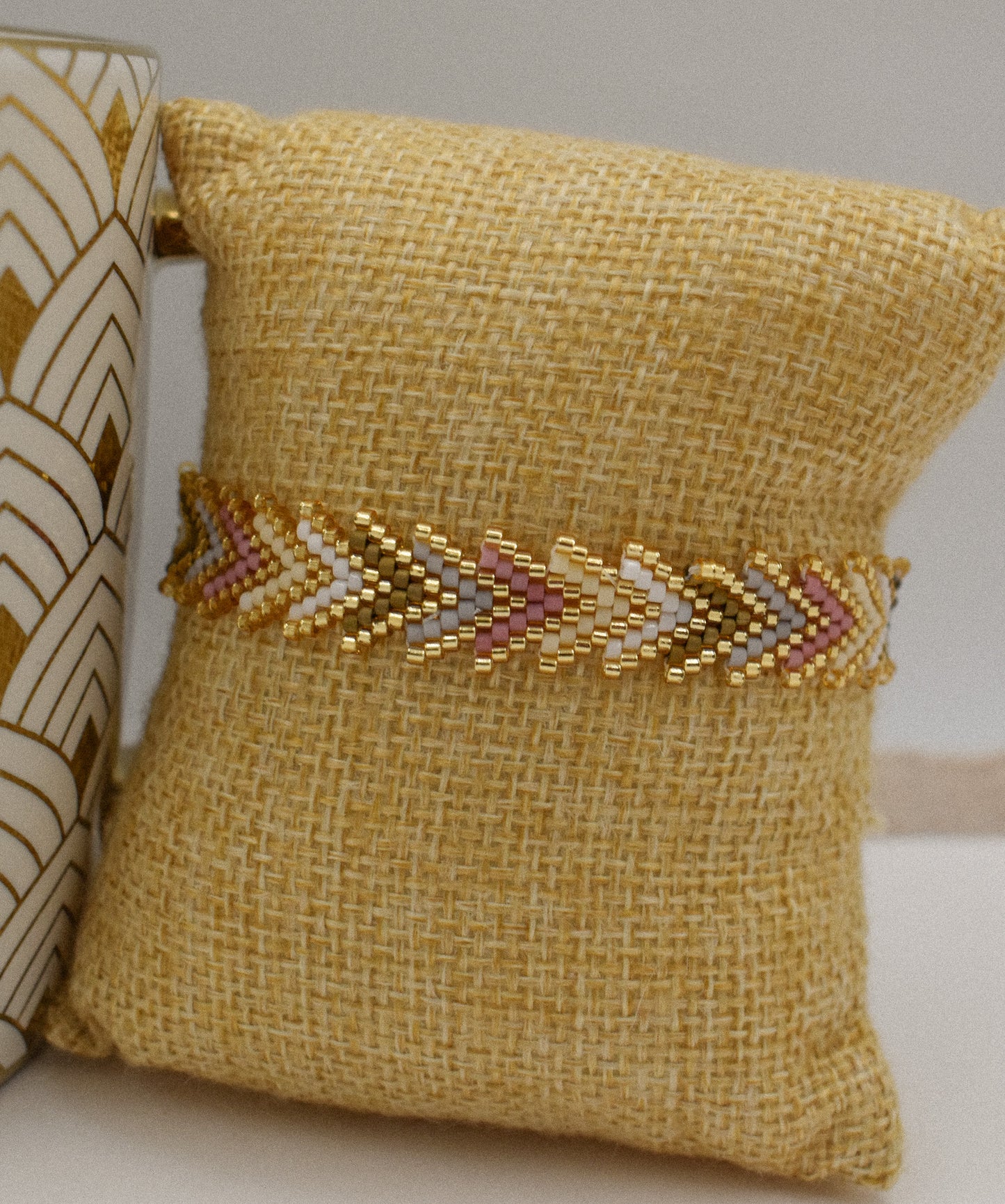 A gold beaded chevron-patterned bracelet displayed on a yellow burlap pillow, with a decorative ceramic vase partially visible on the left.