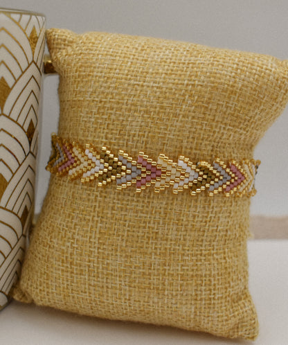 A gold beaded chevron-patterned bracelet displayed on a yellow burlap pillow, with a decorative ceramic vase partially visible on the left.
