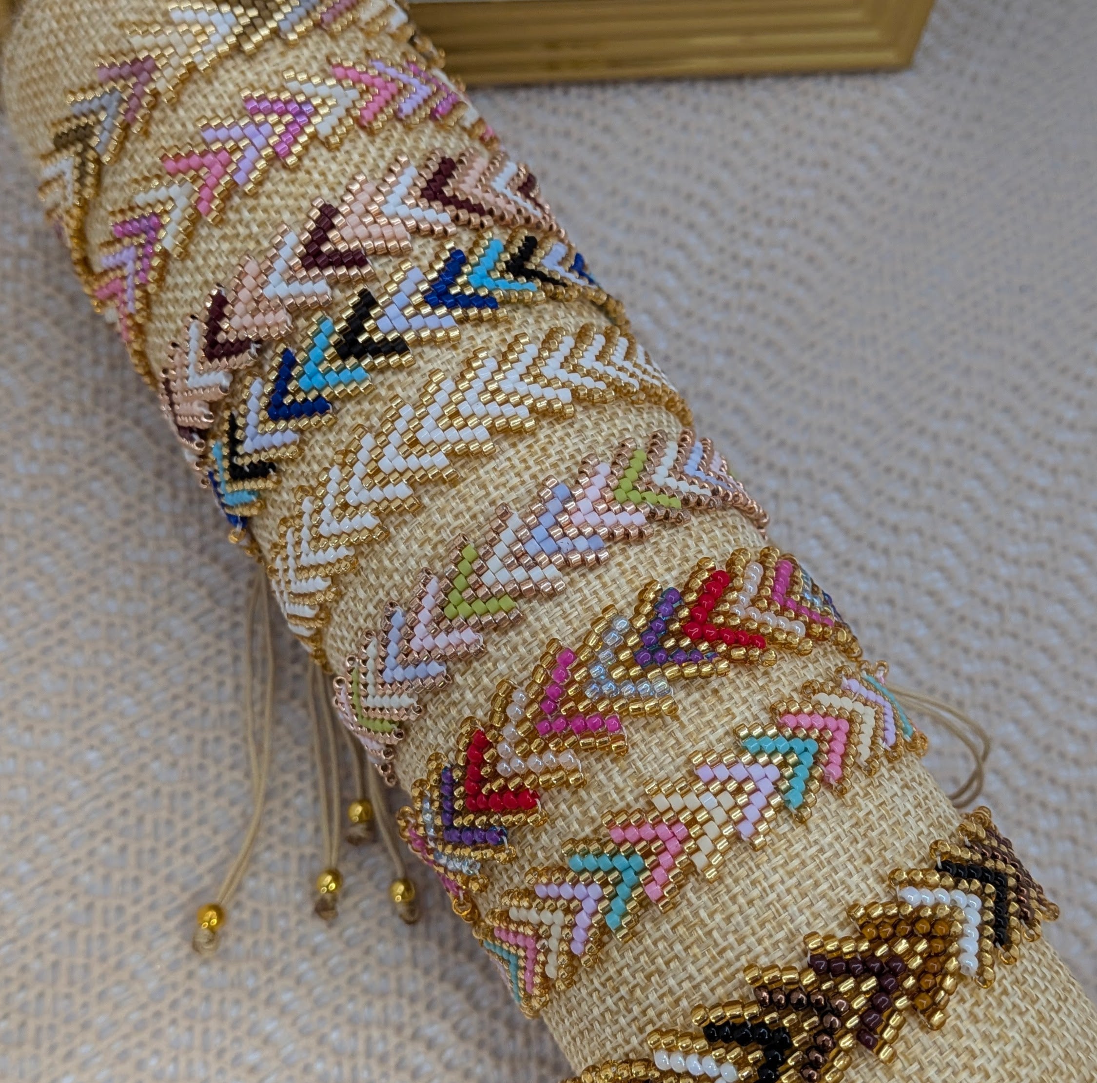 A rolled beige woven textile or basketry piece with colorful chevron beadwork in pink, blue, brown, white, and gold, photographed on a textured light surface with part of a gold-framed object visible in the background.