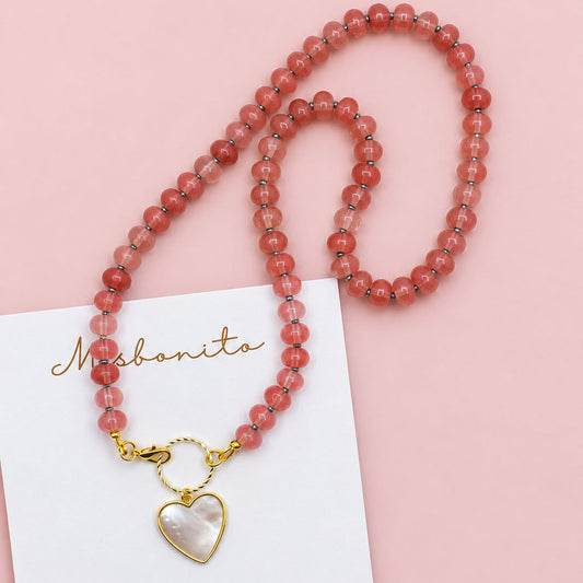 A coral pink beaded necklace with a gold clasp and a white mother-of-pearl heart pendant, displayed on a white Misbonito brand card over a textured beige background.