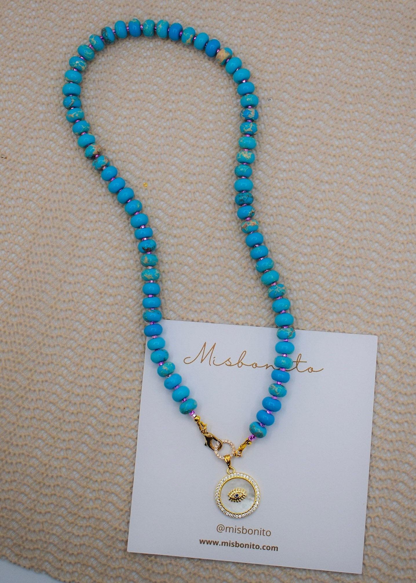 A blue jasper beaded necklace with a gold-toned circular pendant featuring a central eye design, displayed on a white card over a beige textured fabric background.