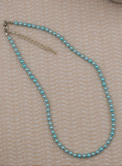 A delicate necklace made of small light blue beads with a silver-tone chain extender, laid on a textured beige fabric background.