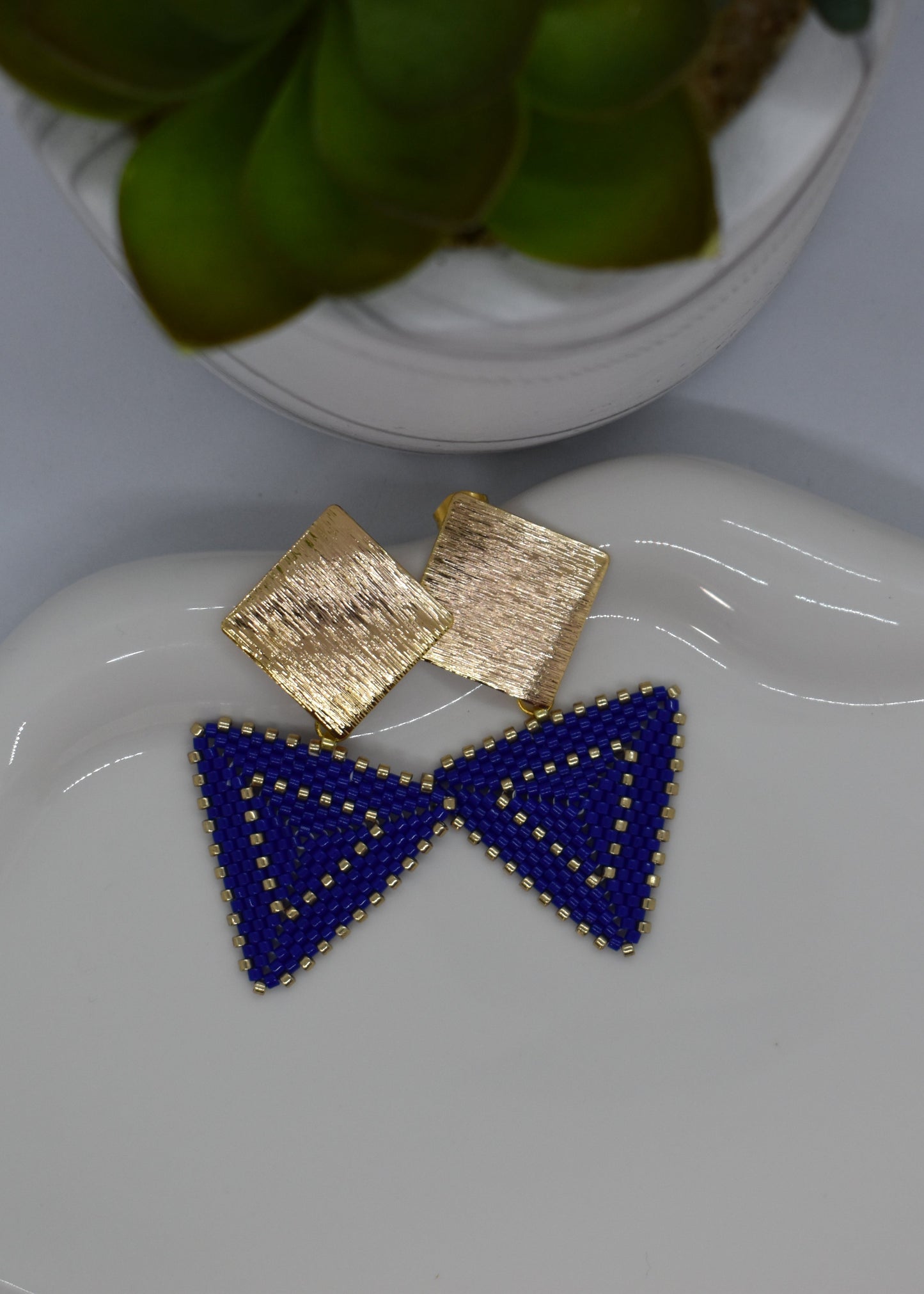 Blue triangle earrings on a white dish with a gold square earring and decorative plate in the background.