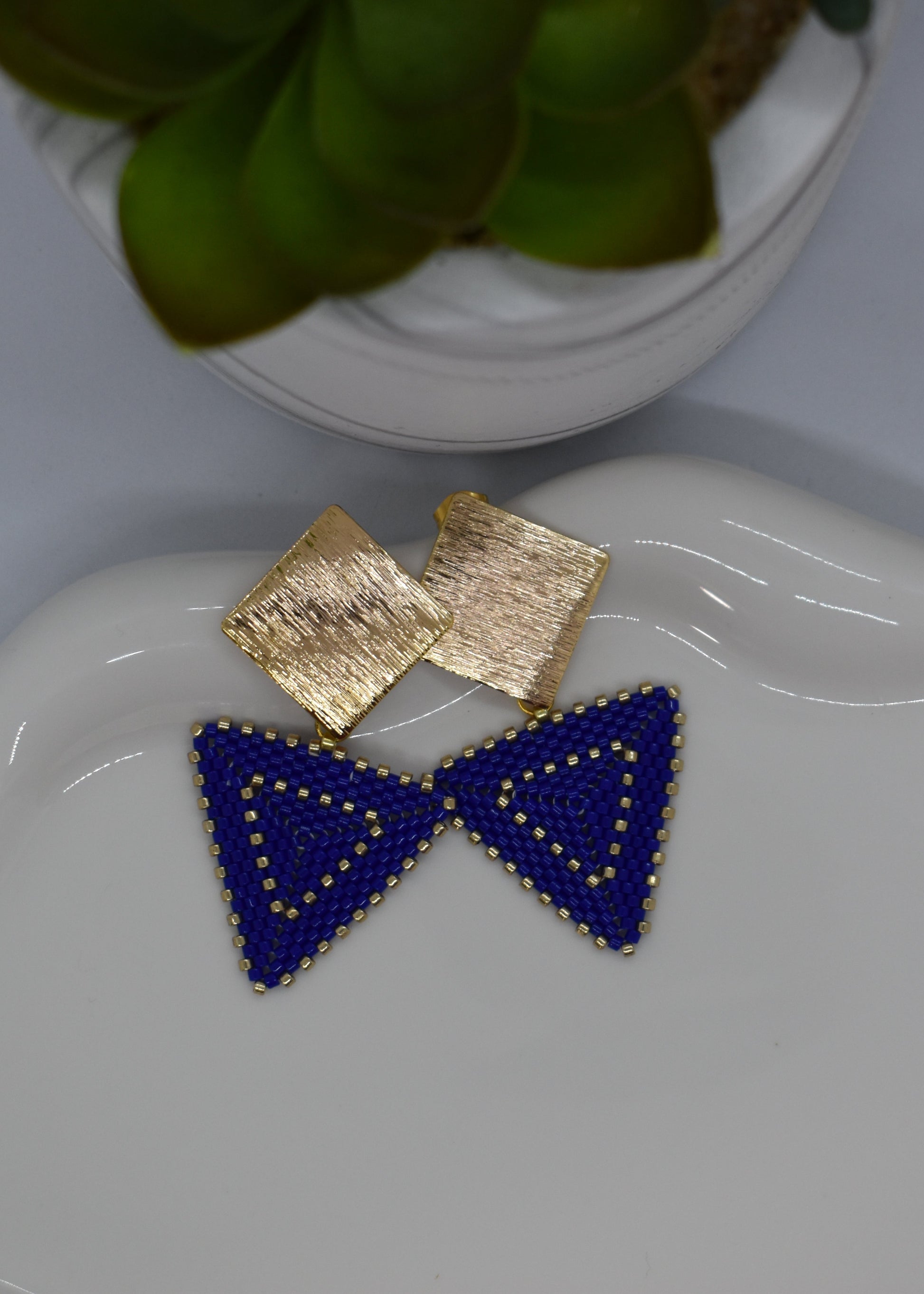 Blue triangle earrings on a white dish with a gold square earring and decorative plate in the background.